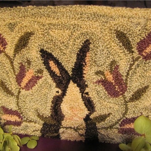 May include: A close-up of a needlepoint rug with a brown rabbit in the center surrounded by purple and pink flowers on a green background.
