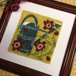 Könnte beinhalten: Ein gerahmtes Nadelbild mit einer blauen Gießkanne mit gelbem Griff und Auslauf, umgeben von braunen und rosa Blumen auf gelbem Hintergrund.