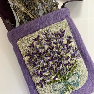 May include: A purple felt bag with a strap, featuring a hand-stitched lavender design. The bag is decorated with a green bow and has a light beige background.