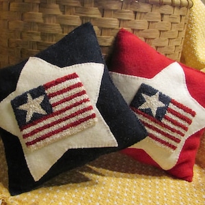 May include: Two decorative throw pillows with a white star and American flag design. The pillows are made of felt and have a navy blue and red background.