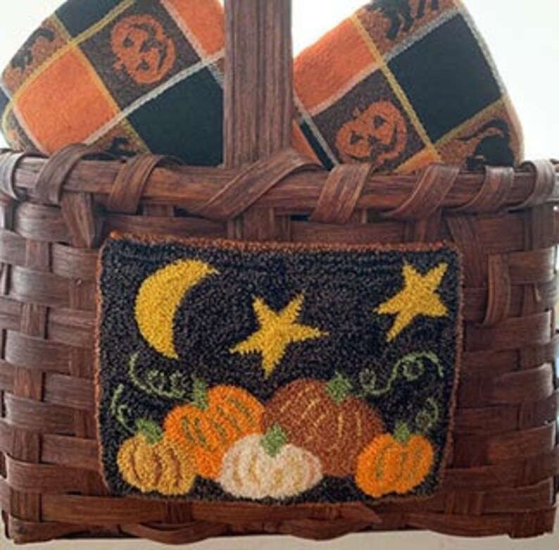 K&ouml;nnte beinhalten: Ein brauner Weidenkorb mit zwei orange-schwarzen karierten Handt&uuml;chern und einem kleinen Nadelbild mit Mond, Sternen und K&uuml;rbissen. Das Nadelbild sagt "Happy Fall".