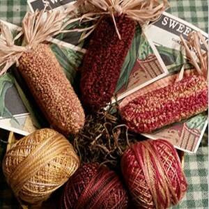 May include: Four yarn balls in shades of red and gold, along with three handmade corn cobs in red and gold, all arranged in a basket with brown straw. The corn cobs are made with yarn and have a rustic, handmade look.