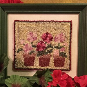 May include: A framed needlepoint artwork depicting three potted pink flowers with green leaves. The flowers are in red pots with a beige background.