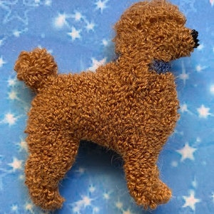 May include: A brown poodle-shaped textile art piece with a blue collar. The poodle has a curly texture and a small, black nose. The background is a light blue with white star shapes.