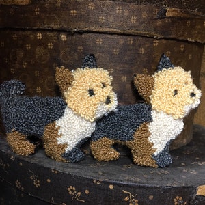 May include: Two small, hand-stitched, plush dog figurines. The dogs are brown, black, and white. They are sitting on a brown, patterned surface.