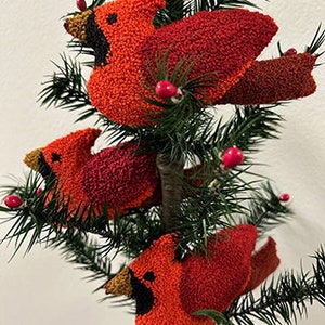 Puede incluir: Tres adornos de pájaros cardenal rojos con ojos y picos negros, y detalles marrones en las alas. Los adornos están colgados de un árbol de Navidad artificial verde.