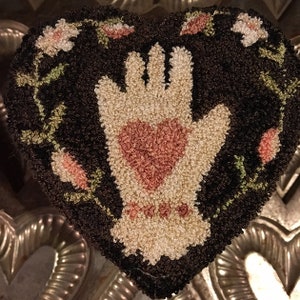 Può includere: Un tappeto marrone a forma di cuore con una mano bianca che tiene un cuore rosa al centro. La mano e il cuore sono circondati da viti verdi e fiori rosa.