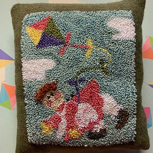 May include: A green felt pillow with a blue felt square in the center. The square features a red, yellow, green, and purple kite flying in the sky with white clouds. A red and white figure is holding onto the kite string.