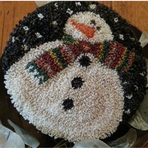 May include: A white snowman with a black hat and a colourful scarf. The snowman has a carrot nose and black eyes. The background is a dark brown wood.