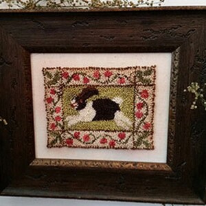 May include: A framed needlepoint design featuring a black and white rabbit running through a green field with red and green flowers. The frame is dark brown wood.