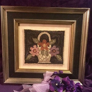 Pode incluir: Uma obra de arte de ponto de agulha enquadrada que representa uma cesta de flores. A cesta é tecida com fio marrom e cheia de flores rosa, roxas e laranja. O fundo é de cor marrom escura.