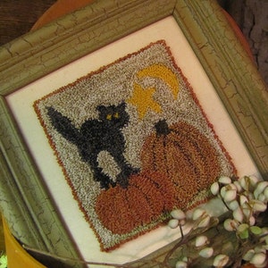 Puede incluir: Una obra de arte de punto de aguja enmarcada que representa un gato negro sentado encima de dos calabazas con una luna creciente en el fondo.