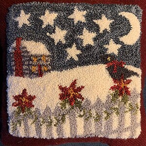 Puede incluir: Una almohada de punto de aguja con una escena de invierno con una pequeña casa, un pájaro negro y flores de poinsettia. El fondo es un cielo azul oscuro con estrellas blancas y una luna creciente.
