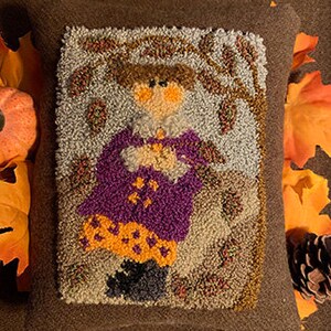 May include: A hand-stitched wool needlepoint pillow featuring a girl in a purple coat and orange and black skirt. The girl is holding a small brown object in her hands. The background is a light blue with brown trees and leaves.