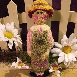 Pode incluir: Uma boneca de feltro feita à mão com um traje rosa e verde, um chapéu amarelo e cabelo castanho. A boneca está em pé em frente a uma cerca branca de madeira e flores brancas.