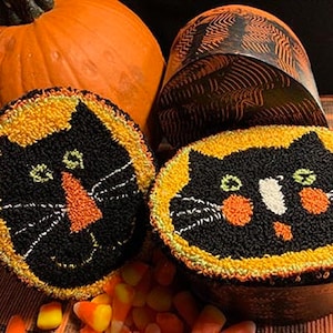 May include: Two black cat needlepoint ornaments with orange and green accents. The ornaments are round and feature a black cat face with a white nose and whiskers. The ornaments are surrounded by candy corn and a pumpkin.