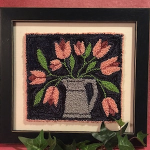Pode incluir: Uma obra de arte de ponto de agulha enquadrada apresentando um buquê de tulipas cor-de-rosa num jarro cinzento sobre um fundo azul escuro.