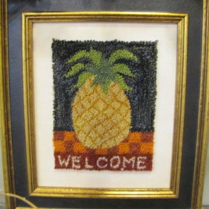 Puede incluir: Una obra de arte de punto de aguja enmarcada que presenta una piña con una parte superior verde y un cuerpo amarillo. La piña está sentada sobre un fondo a cuadros rojo y naranja. El texto "WELCOME" está cosido en blanco debajo de la piña.