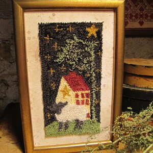 Op de afbeelding: Een ingelijst naaldkunstwerk dat een wit schaap voorstelt dat voor een huis met een rood dak staat met een gele ster aan de zijkant. Het huis is omgeven door groen gras en een hoge boom met een donkerbruine stam. De achtergrond is een donkerblauwe lucht met gouden sterren.