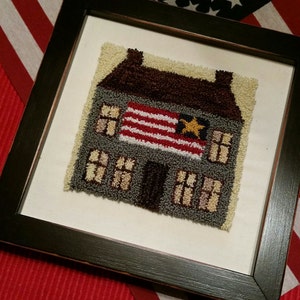 May include: A framed needlepoint artwork depicting a gray house with a brown roof and a red, white, and gold flag on the front. The house has windows and a door. The artwork is on a white background.