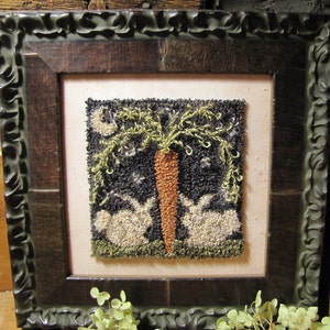 Può includere: Un'opera d'arte in punto ago incorniciata che raffigura due conigli bianchi sotto un albero verde frondoso con una carota al centro. Lo sfondo è nero con stelle bianche. La cornice è marrone scuro con un bordo decorativo e ornato.
