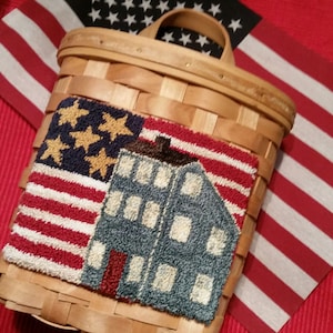 Op de afbeelding: Een geweven mand met een handvat, met een rood, wit en blauw naaldpuntontwerp van een huis met een vlag. Het huis is blauw met witte ramen en een rode deur. De vlag is een traditionele Amerikaanse vlag met rode en witte strepen en witte sterren op een blauw veld.