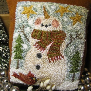 May include: A brown felt pillow with a hand-stitched design of a snowman wearing a red and green scarf. The snowman is surrounded by white snowflakes and yellow stars.