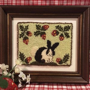 May include: A framed needlepoint design featuring a black and white rabbit in a field of green leaves and red strawberries.
