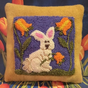 Pode incluir: Uma almofada de feltro amarelo com um desenho bordado quadrado de um coelho branco sentado em um campo de flores roxas. As flores são laranja e amarelas com caules e folhas verdes.