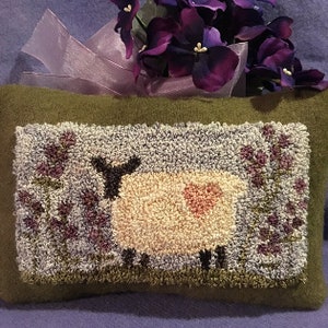 May include: A green wool pillow with a hand-stitched design of a white sheep with a pink heart on its side, standing in a field of purple flowers.