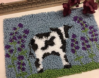 May include: A framed needlepoint design featuring a black and white cow standing in a field of purple flowers. The background is a light blue color.