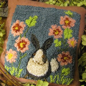 Pode incluir: Uma almofada de ponto de agulha com um coelho preto e branco rodeado de flores cor-de-rosa e amarelas sobre um fundo azul.