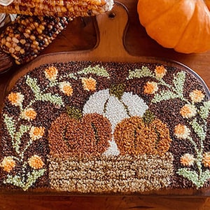 May include: A wooden craft hoop with a needlepoint design featuring three pumpkins, two orange and one white, with green leaves and yellow flowers. The pumpkins are in a brown basket with a wooden handle.