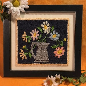 May include: A framed needlepoint artwork featuring a bouquet of colorful flowers in a gray pitcher on a dark blue background.
