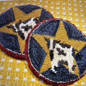 May include: Two round, hand-tufted coasters. Each coaster features a gold star on a navy blue background, with a cow silhouette in the center. The coasters have a red border and are set on a yellow patterned surface.