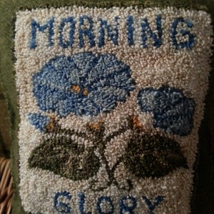 May include: A green fabric pillow with a white background and blue embroidery. The embroidery depicts a morning glory flower with the words "Morning Glory" above the flower and "Glory" below the flower.