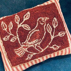 Puede incluir: Una pequeña almohada cuadrada roja con un diseño bordado blanco de un pájaro posado en una rama con hojas.