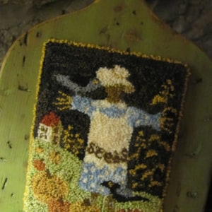 May include: A colourful rug hooking depicting a scarecrow with a white shirt, blue trousers, and a black hat. The scarecrow is standing in a field with pumpkins, sunflowers, and a small house. The rug hooking is framed on a green wooden board.