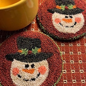 May include: Two round red felt coasters with a snowman design. Each coaster features a white snowman face with black eyes, a carrot nose, and a black top hat with green holly.