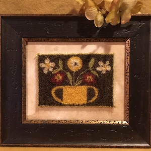 May include: A framed needlepoint artwork featuring a yellow cup with a brown handle filled with three white flowers and two red flowers on a dark green background.