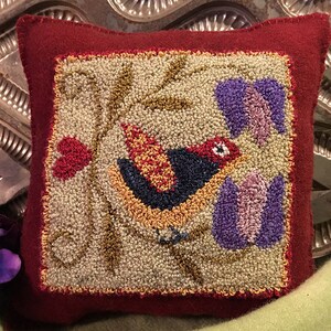 Puede incluir: Una almohada roja con un cuadrado de color crema en el centro. El cuadrado presenta un pájaro colorido con acentos rojos, amarillos, naranjas y azules, posado en una rama con hojas verdes y flores moradas.