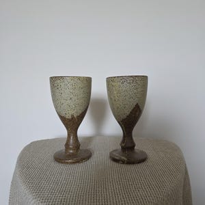 May include: Two ceramic goblets featuring a speckled light beige upper section and a solid dark brown base. The vessels have a rounded bowl, a slender stem, and a circular foot, showcasing a distinct two-tone color transition with a textured finish.
