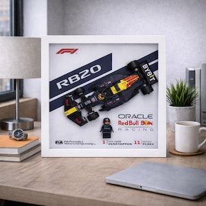 Può includere: Opera d'arte incorniciata con un modello di auto da corsa Red Bull RB20 e una figura di pilota in miniatura. L'opera include il logo Oracle Red Bull Racing, i nomi dei piloti e i dettagli del Campionato del Mondo FIA di Formula 1. La cornice è bianca.