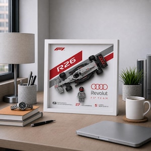 May include: A framed image of a white and grey R26 race car model with red detailing. The frame displays the Audi Revolut F1 Team logo and driver information. A small Lego figure is also included. The image is on a wooden desk.