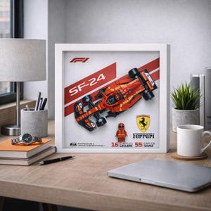 Puede incluir: Un modelo LEGO enmarcado de un coche de carreras Ferrari SF-24 de Fórmula 1 rojo y negro. El marco incluye el logotipo de Ferrari, los nombres de los pilotos y el texto "SF-24". También se incluye una pequeña figura LEGO. El marco es blanco.