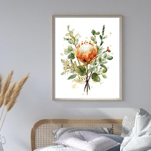 Puede incluir: Impresión en acuarela de una flor de protea con pétalos naranjas y blancos, rodeada de hojas verdes y follaje. La obra de arte está enmarcada en un marco de madera clara y se exhibe en un dormitorio. La habitación también cuenta con un jarrón blanco con plantas secas.