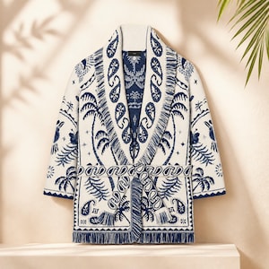 May include: A cream-colored knit cardigan featuring a dark blue paisley and palm tree pattern. The garment includes a shawl collar, a matching tie belt, and fringe detailing along the front edges, cuffs, and hemline, creating a textured look.