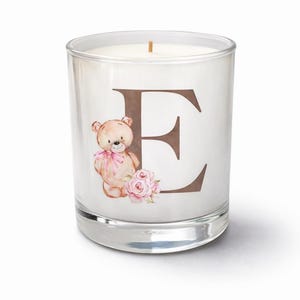May include: A clear glass candle with a brown letter "E" and a teddy bear illustration. The teddy bear has a pink bow and is next to a bouquet of pink roses. The candle is a decorative item.