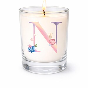 May include: A lit candle in a clear glass jar. The candle has a white wax base with a large letter "N" in a gradient of pink and purple, decorated with seashell illustrations. The candle is a decorative item.