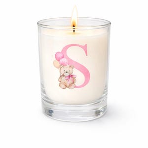 May include: A lit candle in a clear glass jar. The candle has a pink letter "S" with a teddy bear holding balloons. The teddy bear has a pink bow. The candle is on a white background.